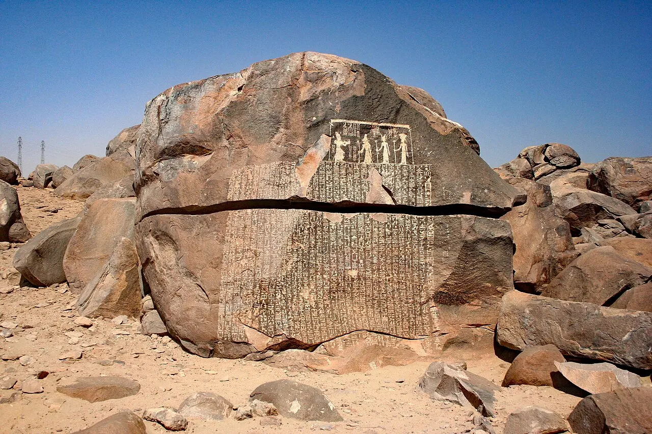 Um grande bloco de granito rachado ao meio, com colunas de hieróglifos gravadas na face e uma cena de figuras no alto, entre outros blocos de pedra num terreno de areia.