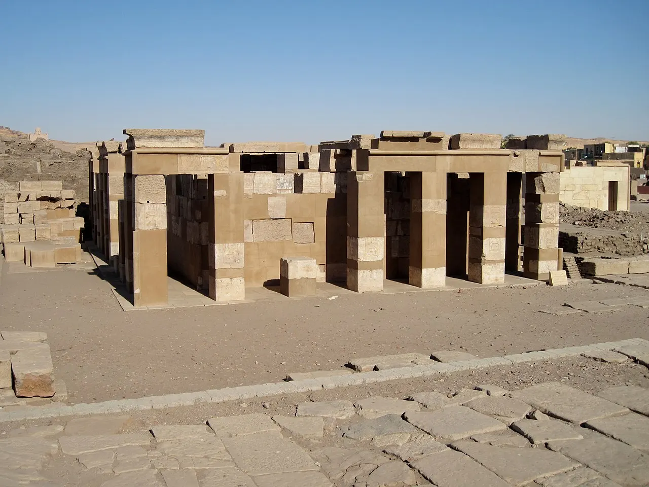 Un pequeño templo de pilares cuadrados de arenisca, sin techo completo, sobre un terreno llano de tierra y losas, con bloques de piedra apilados a la izquierda y cielo azul.