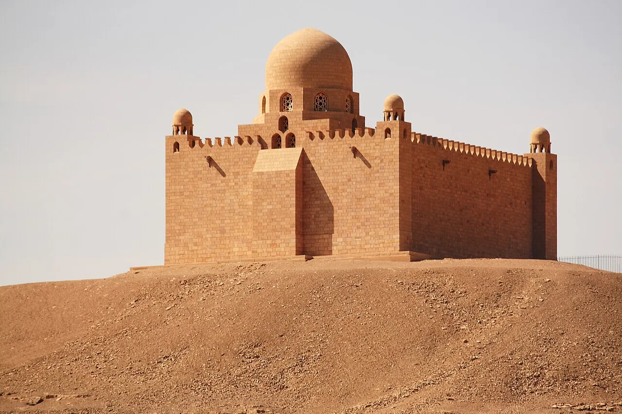 Um edifício de pedra rosada com cúpula central, pequenas cúpulas nos cantos e ameias no alto dos muros, no topo de uma colina de areia, sob céu claro.