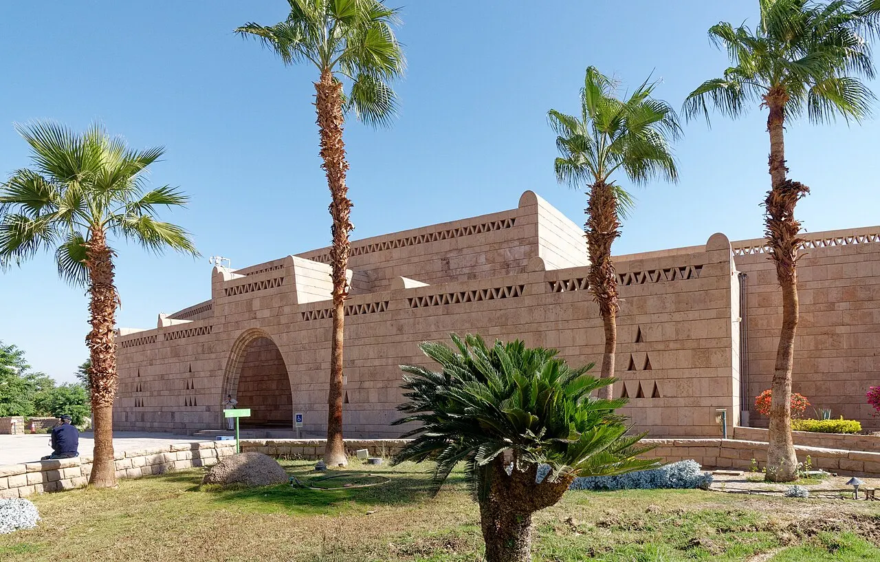 The pinkish stone facade of the Nubian Museum, with an arched entrance and friezes of triangular openings, seen from the garden with lawn, tall palms and a cycad in the foreground.