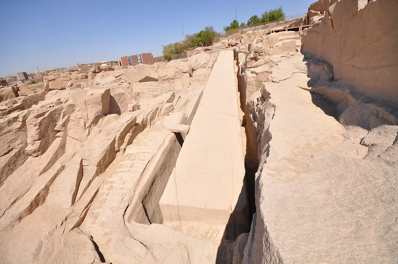 A long block of pinkish granite in the shape of an obelisk, still attached to the bedrock, between trenches cut into the quarry, with city buildings in the background.
