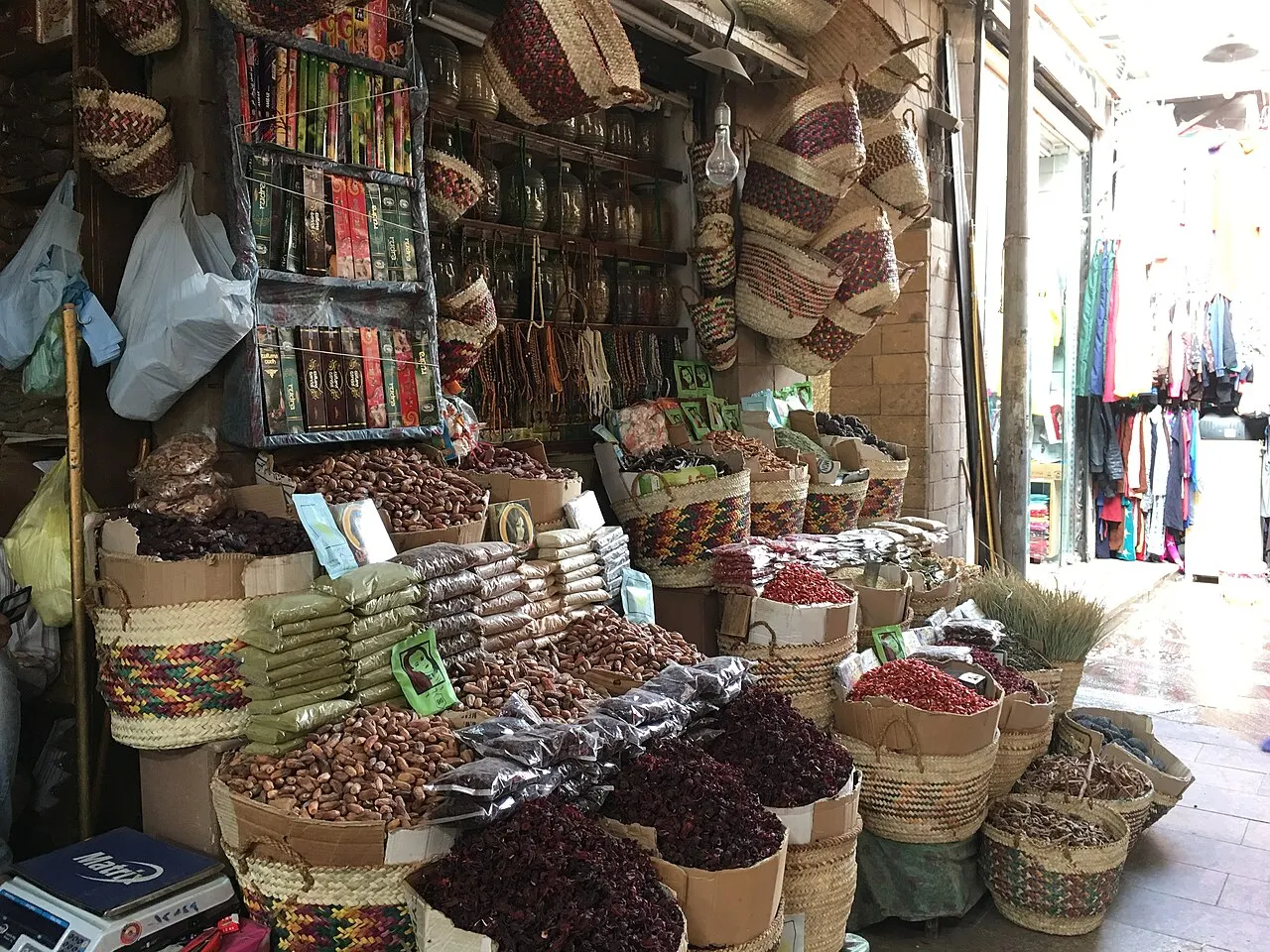 Un puesto de mercado con cestos de paja llenos de dátiles, flores secas rojas y granos, paquetes apilados, estantes con frascos e inciensos y cestos colgados encima.