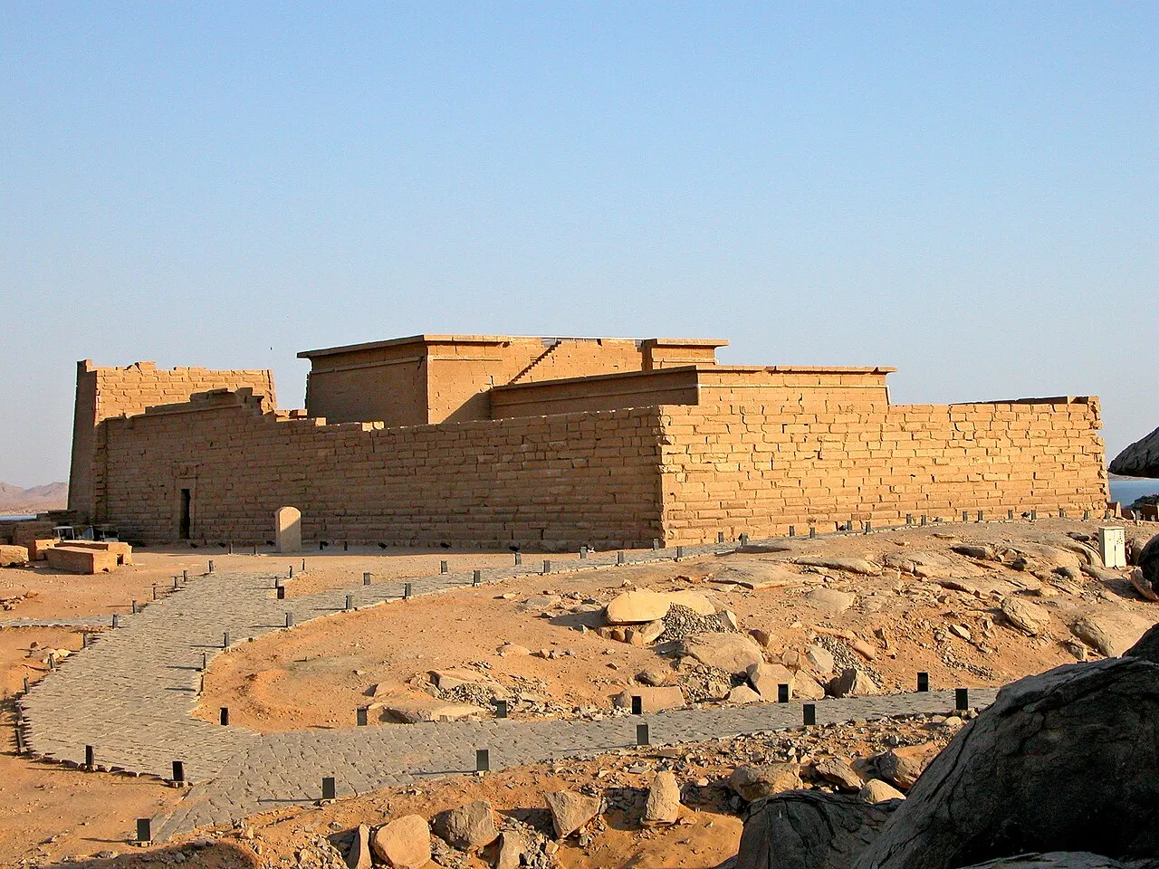 El templo de arenisca de muros altos y lisos, iluminado por el sol bajo, sobre un terreno de rocas con un camino de piedras bordeado por pequeños postes.