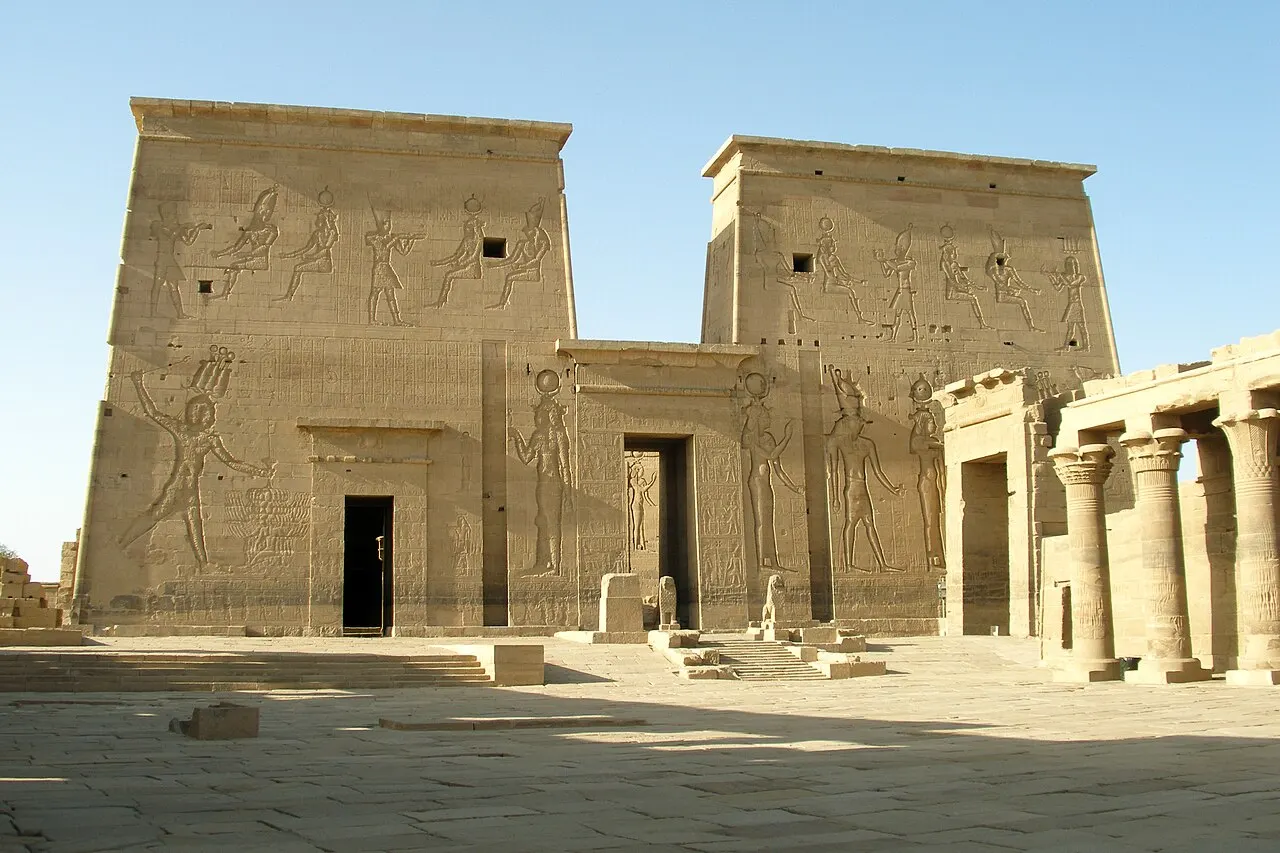 El primer pilono del templo de Isis en Filé, dos torres de piedra con figuras grabadas en relieve y una puerta baja en el centro, con una hilera de columnas a la derecha y el patio enlosado delante, bajo un cielo azul.