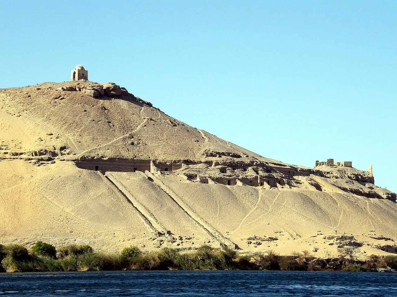 A sandy hill across the river, with a row of entrances cut into the rock halfway up, ramps running down to the water and a small dome on the summit, under a blue sky.
