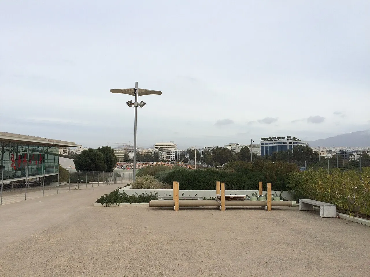 Terraço pavimentado do centro cultural, com a fachada de vidro à esquerda, bancos e a cidade ao fundo.