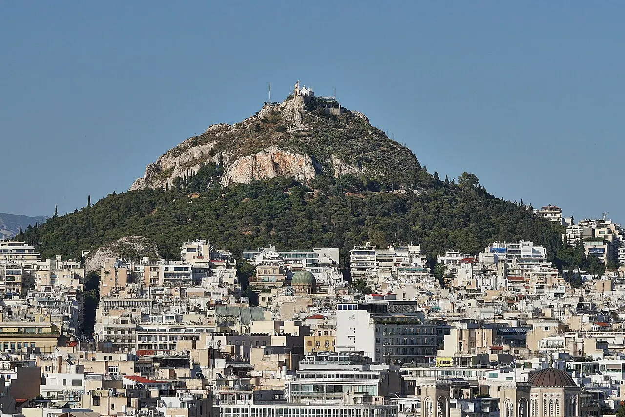 O cone rochoso do monte Licabeto coberto de pinheiros, com a capela branca de São Jorge no cume, erguendo-se acima do mar de prédios baixos e brancos de Atenas.