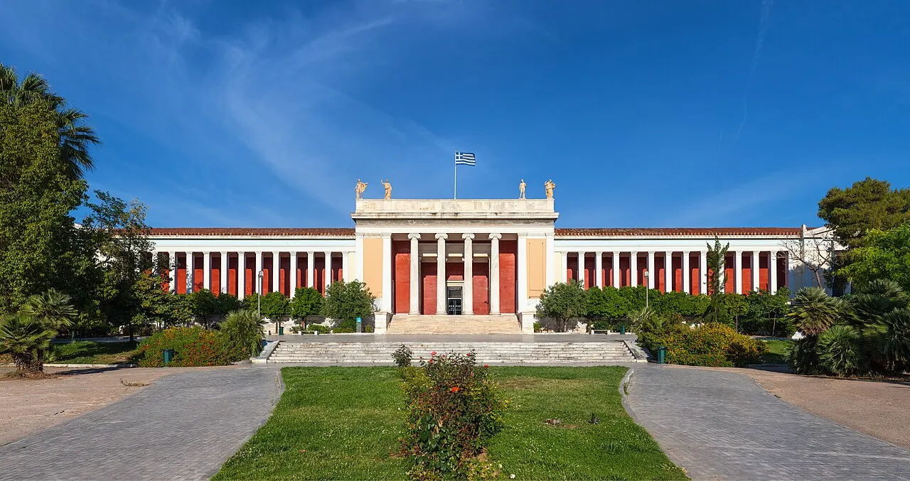 Fachada neoclássica do Museu Arqueológico Nacional de Atenas, com colunas e detalhes vermelhos, atrás do jardim.