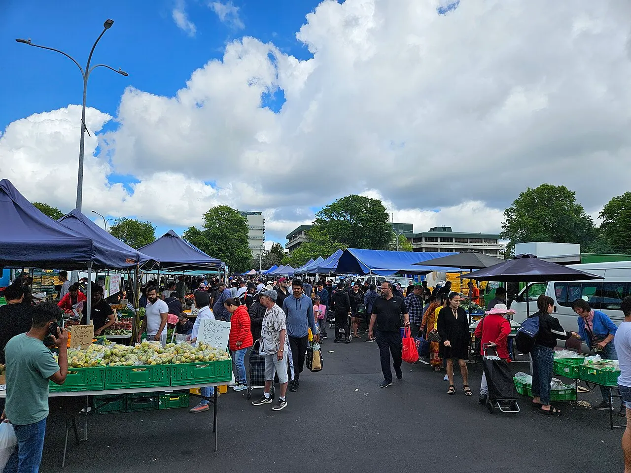 Barracas com toldos azuis armadas num estacionamento, com vendedores e compradores circulando entre caixas de frutas e verduras sob um céu parcialmente nublado.