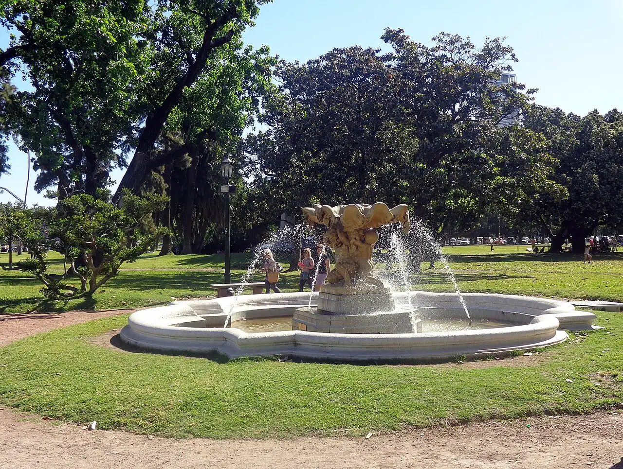 Fonte com jatos de água no gramado dos Bosques de Palermo, com árvores grandes atrás.
