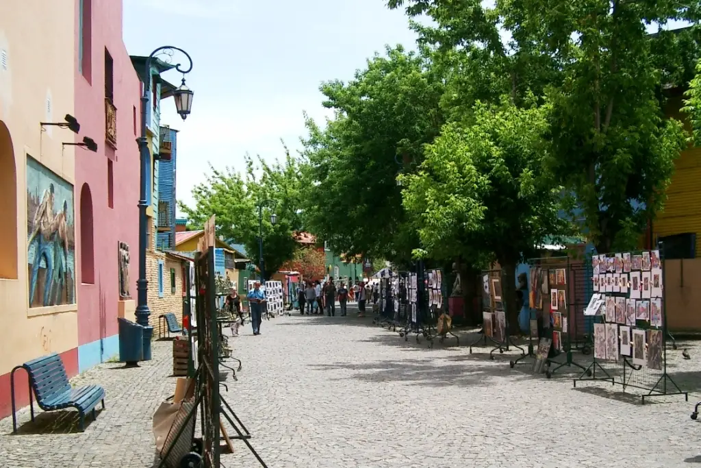 Rua de paralelepípedos do Caminito, com casas coloridas, árvores e quadros expostos nas calçadas.