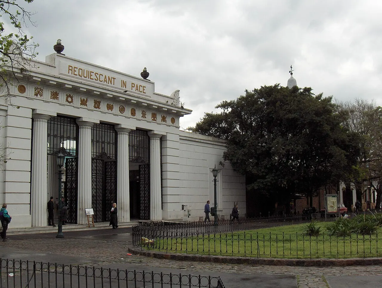 A entrada de um cemitério em estilo neoclássico, com colunas brancas, portões de ferro e a inscrição "Requiescant in Pace" no friso, pessoas passando pela calçada sob céu nublado.