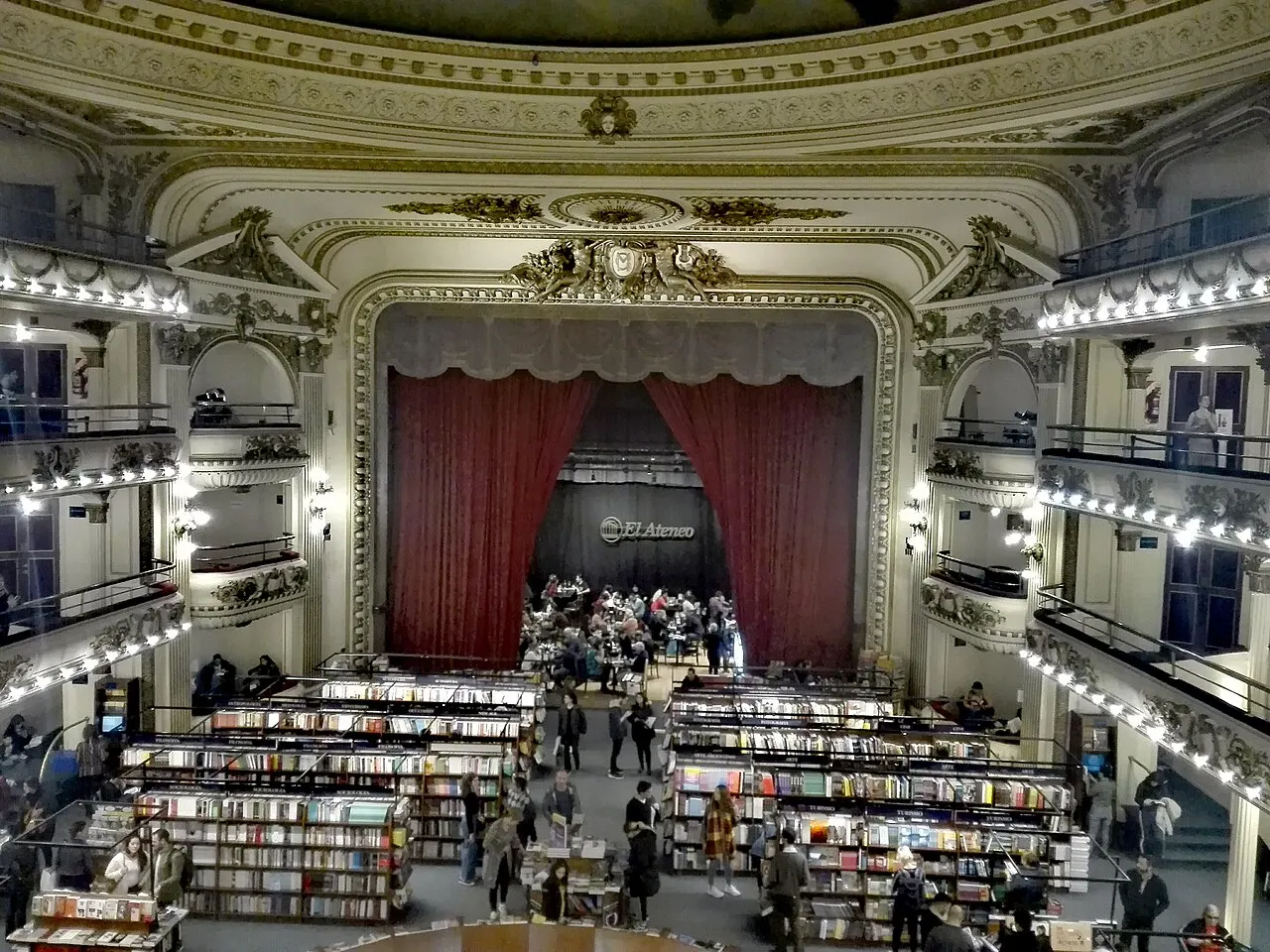 Salão do teatro com a cortina vermelha e estantes de livros no lugar da plateia.