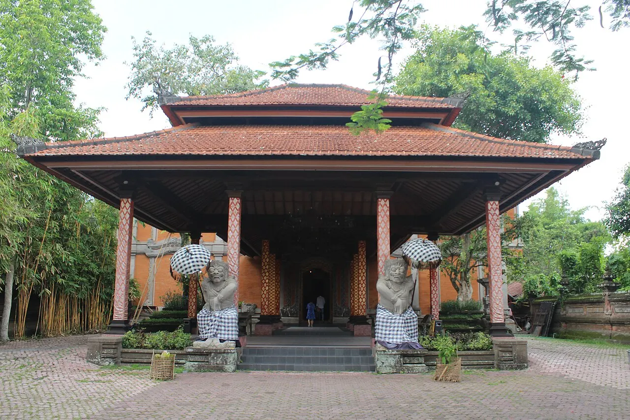 Pavilhão aberto com telhado de telhas terracota, colunas de tijolo e duas estátuas guardiãs vestidas de pano.