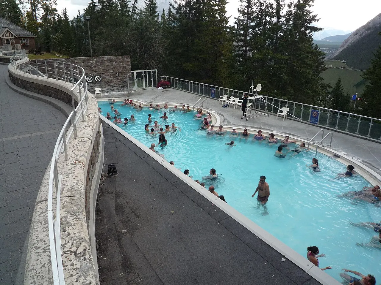 Piscina ao ar livre de água azul-clara com banhistas, cercada por um muro curvo de pedra e um terraço com cadeiras brancas, com pinheiros e montanha atrás.