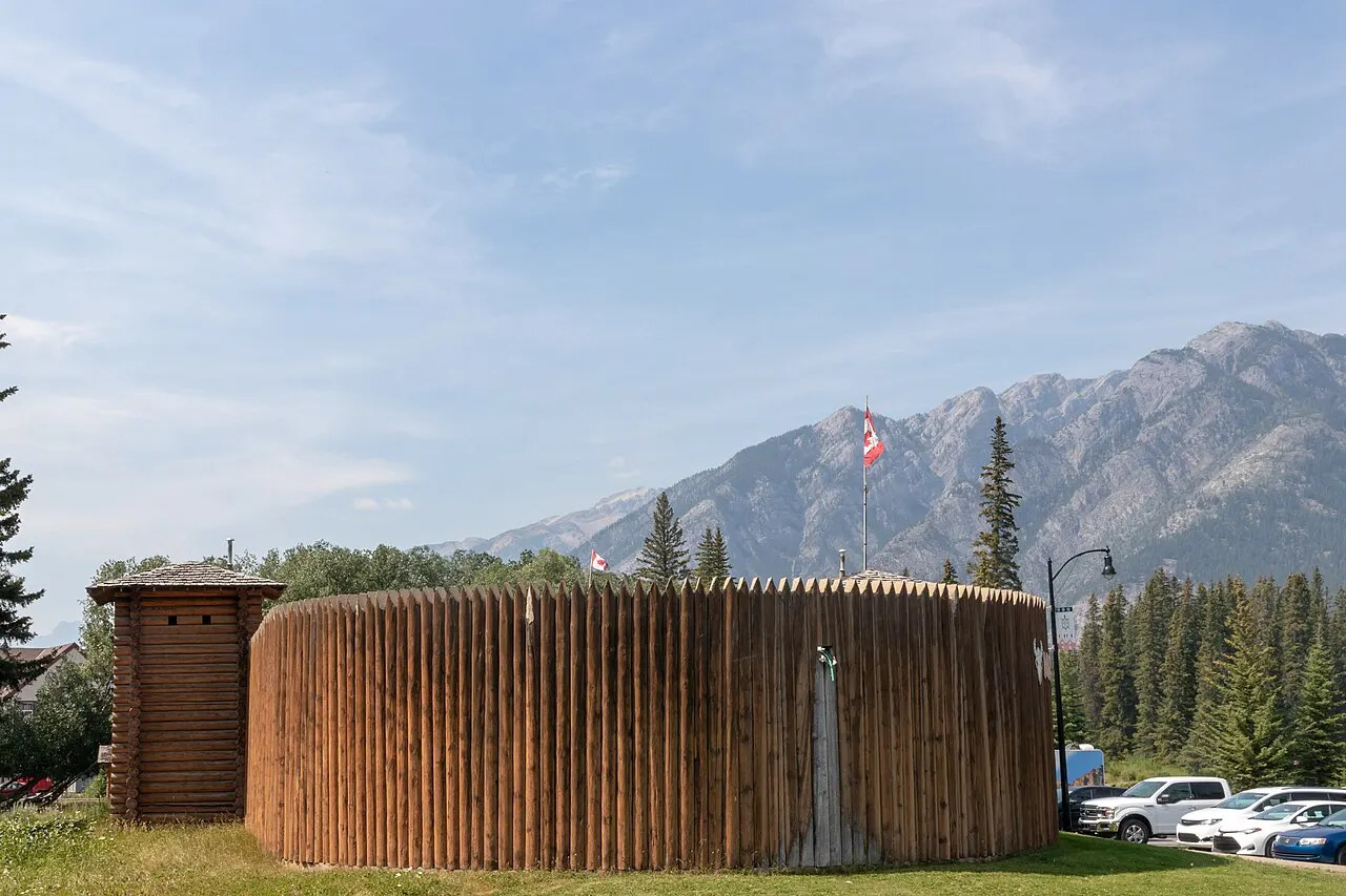 Muro circular de troncos pontudos, como um forte, com uma torre quadrada de madeira ao lado e uma bandeira do Canadá, diante de montanhas cinzentas.