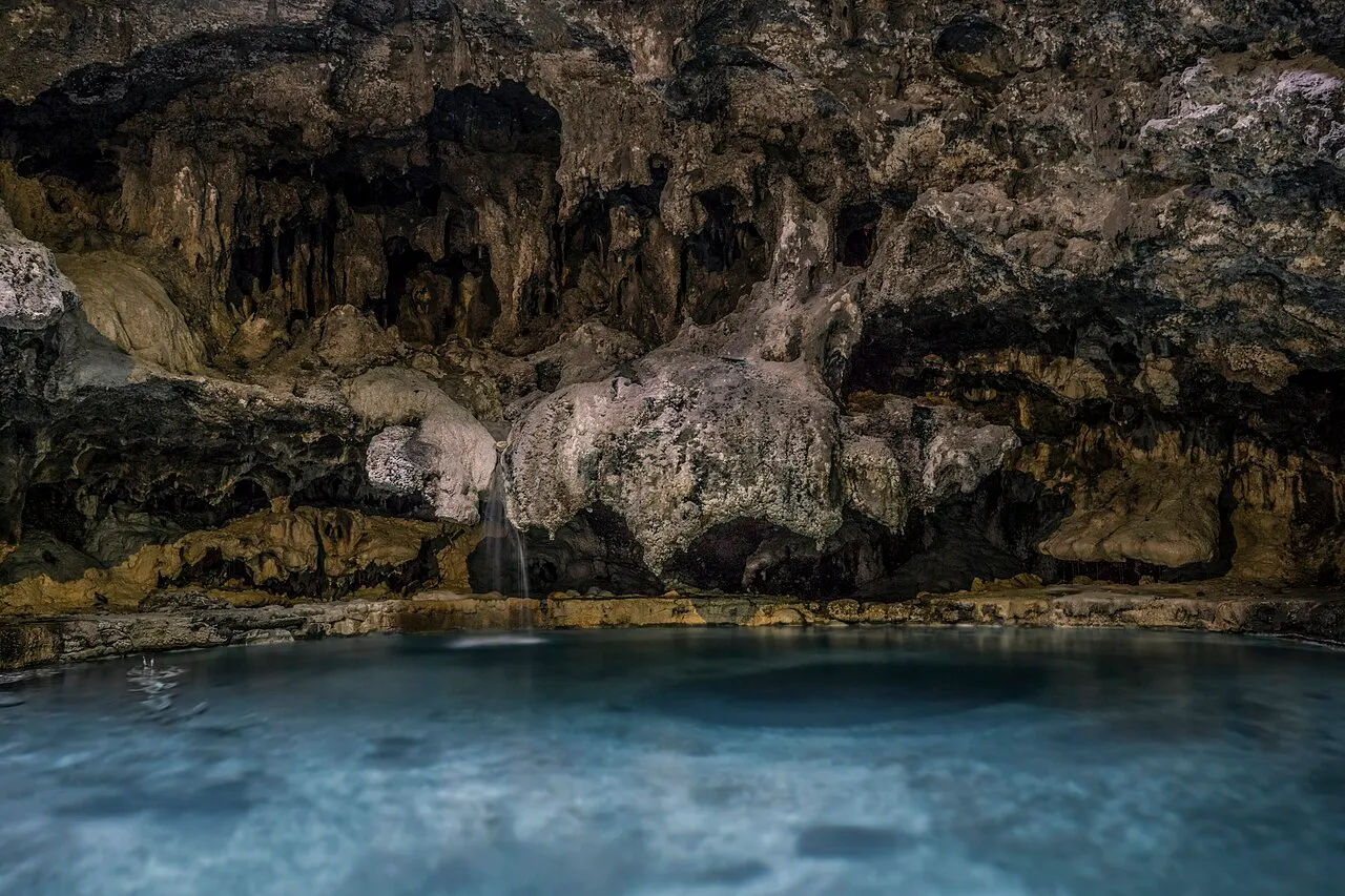 Interior de uma caverna de rocha escura e manchada, com um fio de água escorrendo pela pedra até uma piscina de água azul.