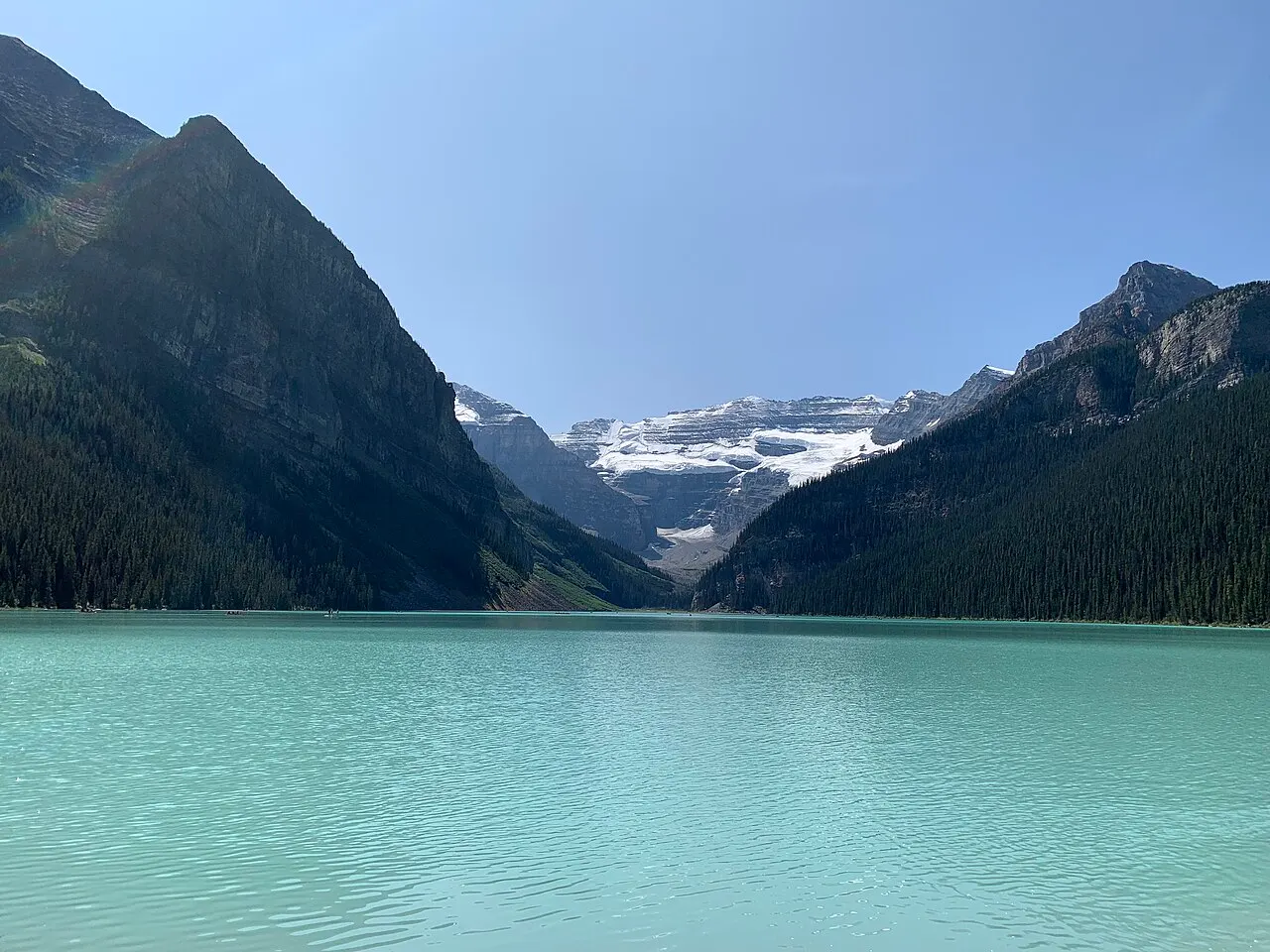 Lago de água verde-clara e lisa entre duas encostas escuras de pinheiros, com uma montanha de geleira branca no fundo do vale, sob céu azul.