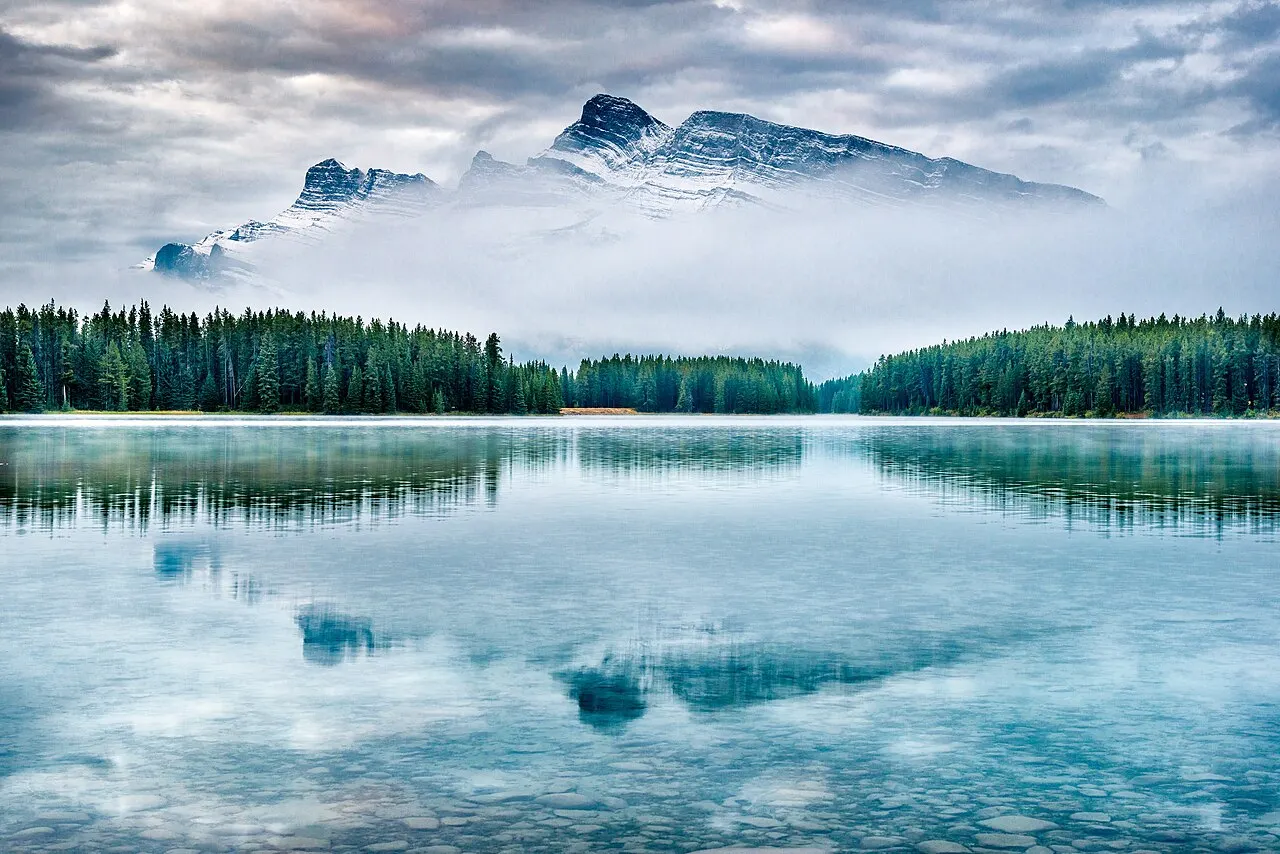 Lago calmo de manhã com névoa sobre a água, uma faixa de pinheiros na margem oposta e o pico de uma montanha aparecendo acima das nuvens, refletido na superfície.