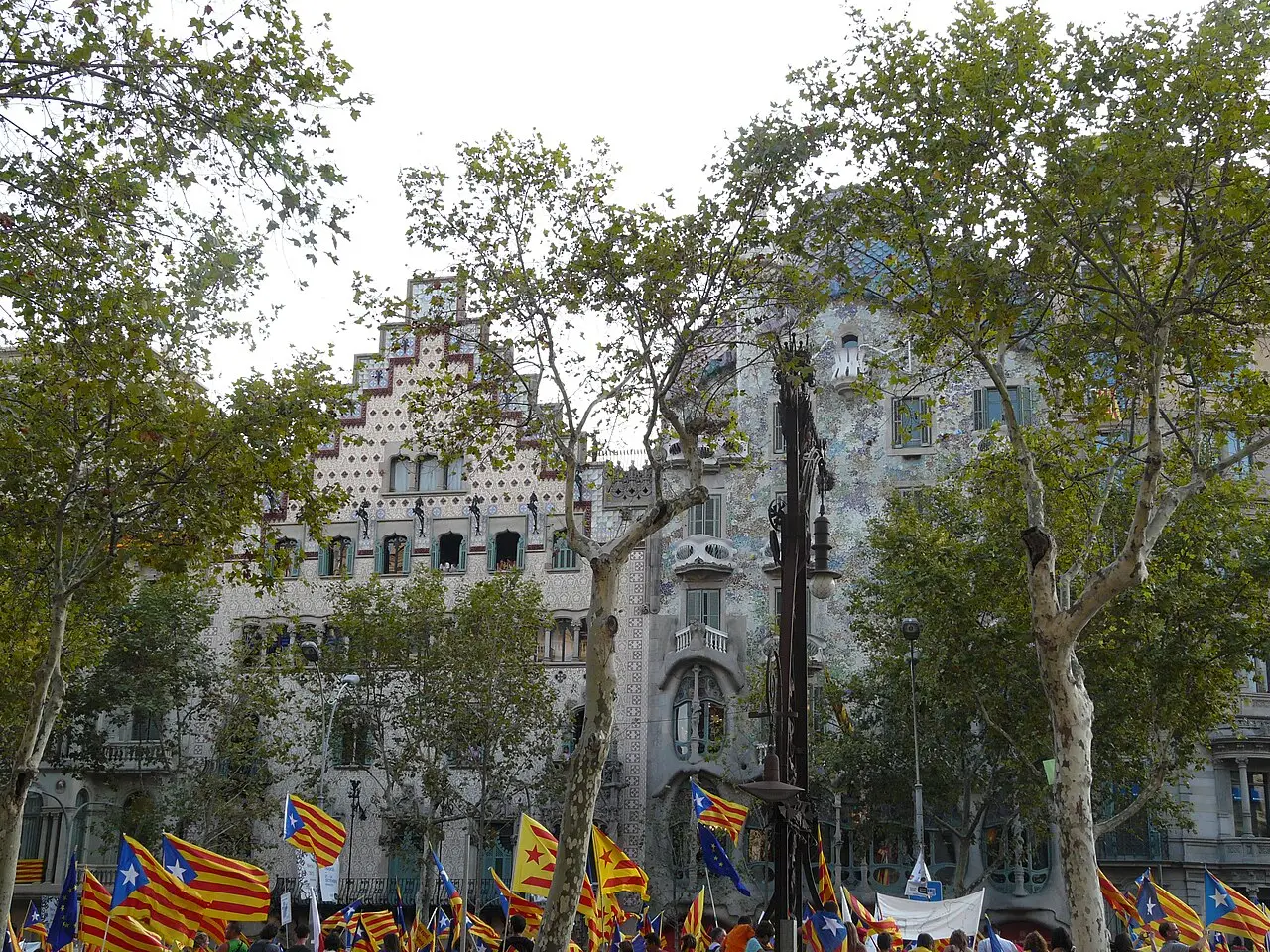 Fachada de pedra da Casa Batlló e o telhado escalonado, com árvores e bandeiras na frente.