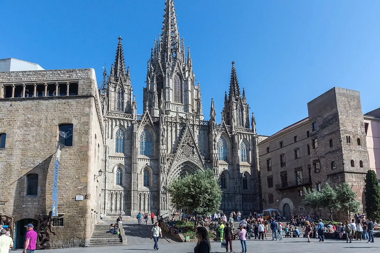 Fachada de pedra com três pináculos da catedral de Barcelona sob céu azul, com a praça em frente ocupada por pedestres e prédios medievais dos dois lados.