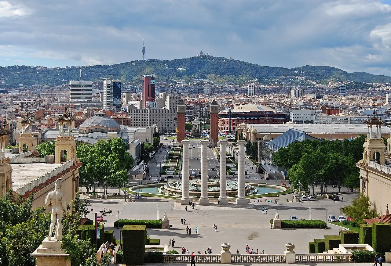 Vista do alto de Montjuïc sobre as quatro colunas, a fonte e a avenida que desce à Plaça Espanya, com as duas torres venezianas e a cidade até a serra de Collserola ao fundo.