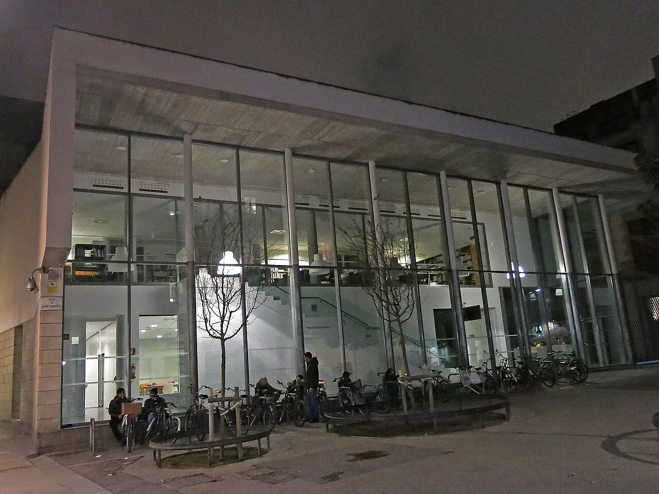 Prédio de vidro do Museu Picasso à noite, com bicicletas na frente e pessoas junto à porta.