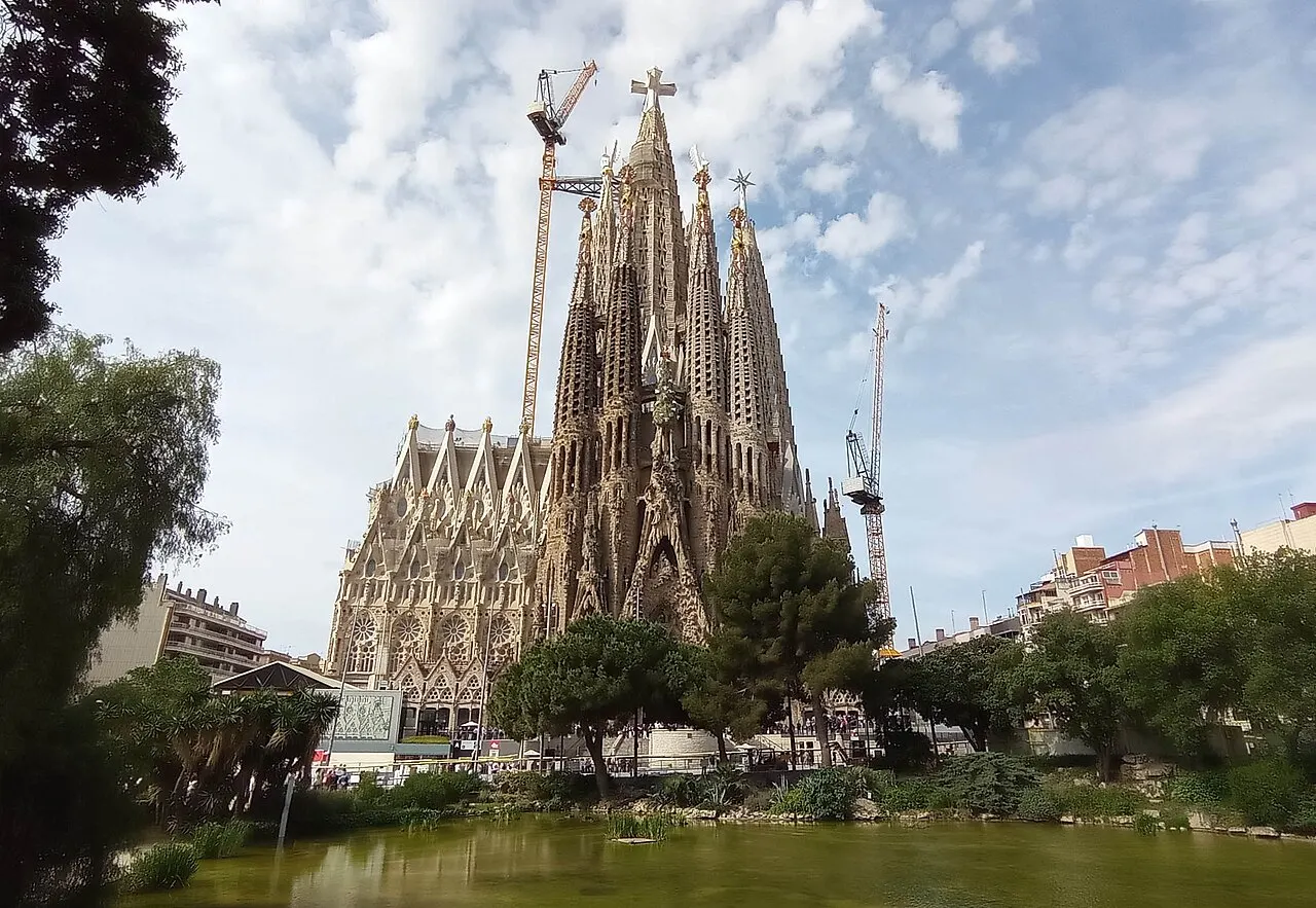 Sagrada Família, em Barcelona, com as torres de pedra e guindastes da obra, atrás das árvores da praça.