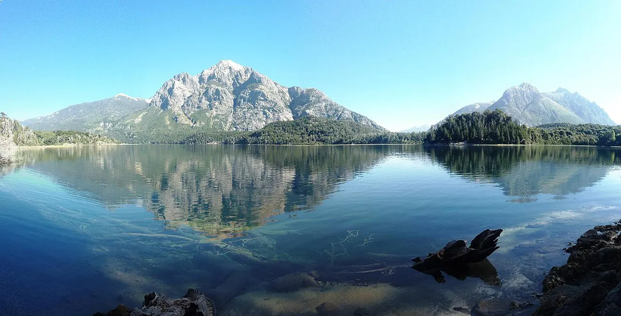 Águas transparentes e paradas do Lago Moreno refletem como espelho as montanhas nevadas ao fundo, com pedras e troncos visíveis sob a superfície.