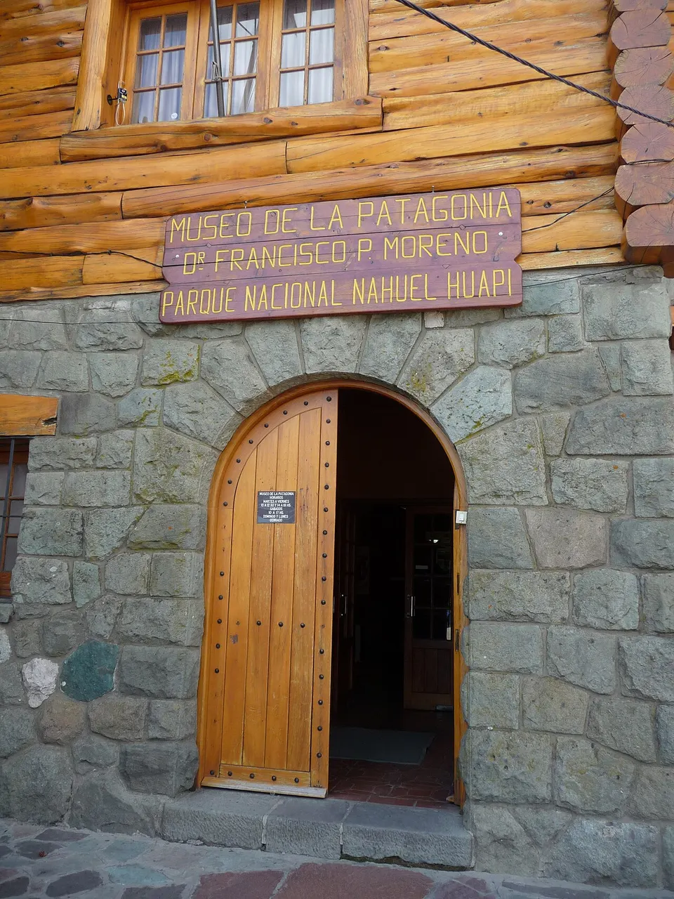 Porta de madeira em arco do museu, na parede de pedra e troncos, com a placa acima.
