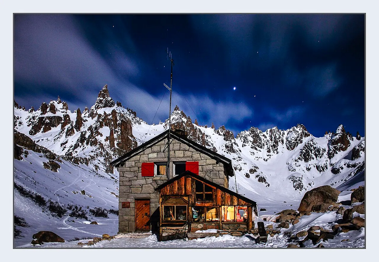 Refúgio Frey, cabana de pedra e madeira com janelas acesas, à noite, entre montanhas com neve.