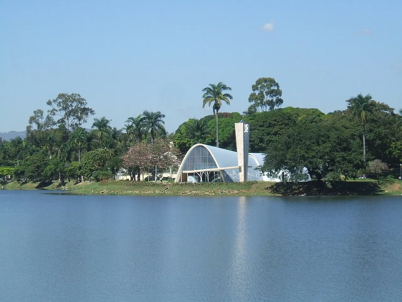 A água calma da Lagoa da Pampulha com a Igrejinha branca e as palmeiras na outra margem.