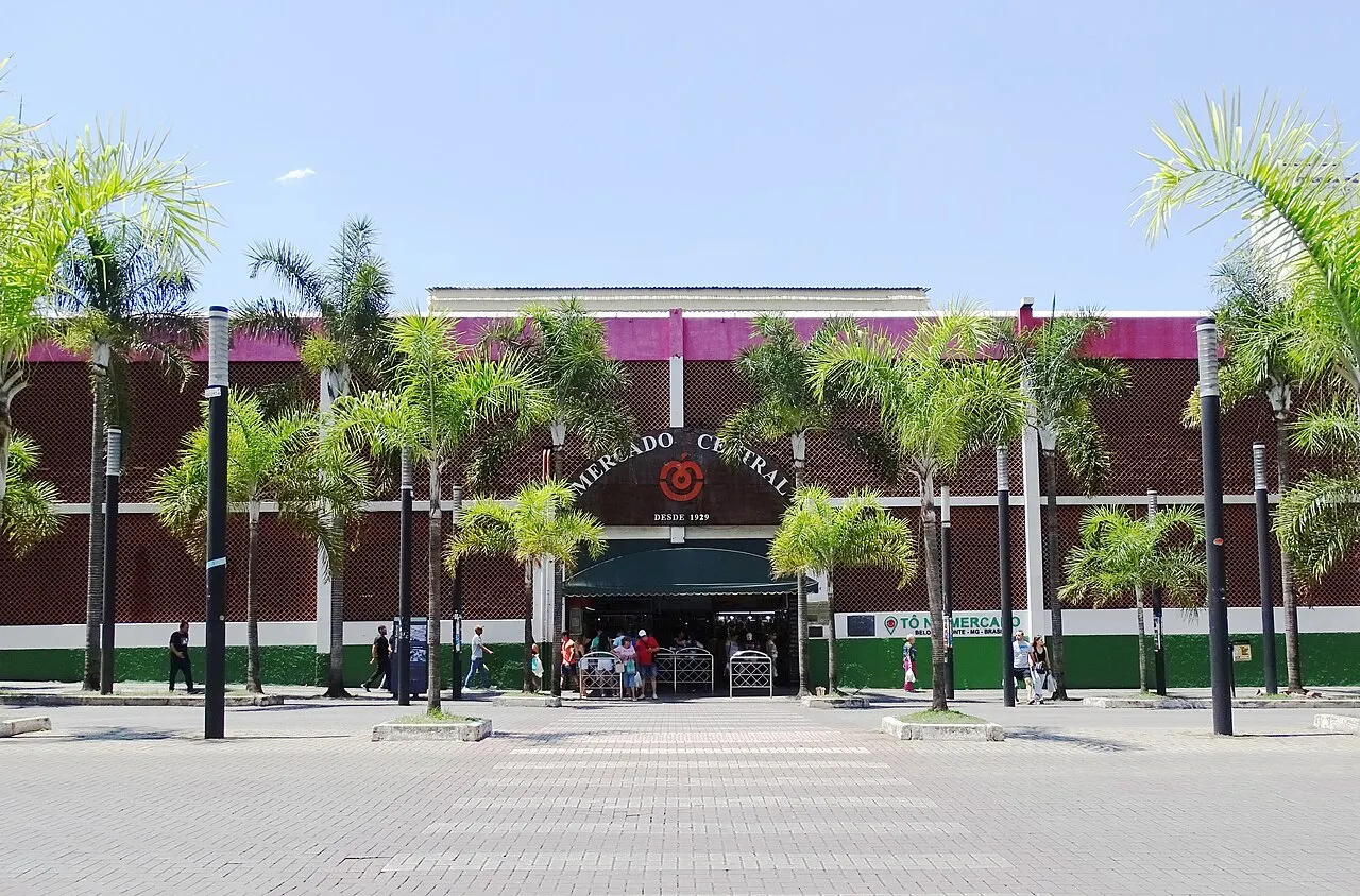 A fachada do Mercado Central de Belo Horizonte vista da praça, entre uma fileira de palmeiras, sob céu limpo.