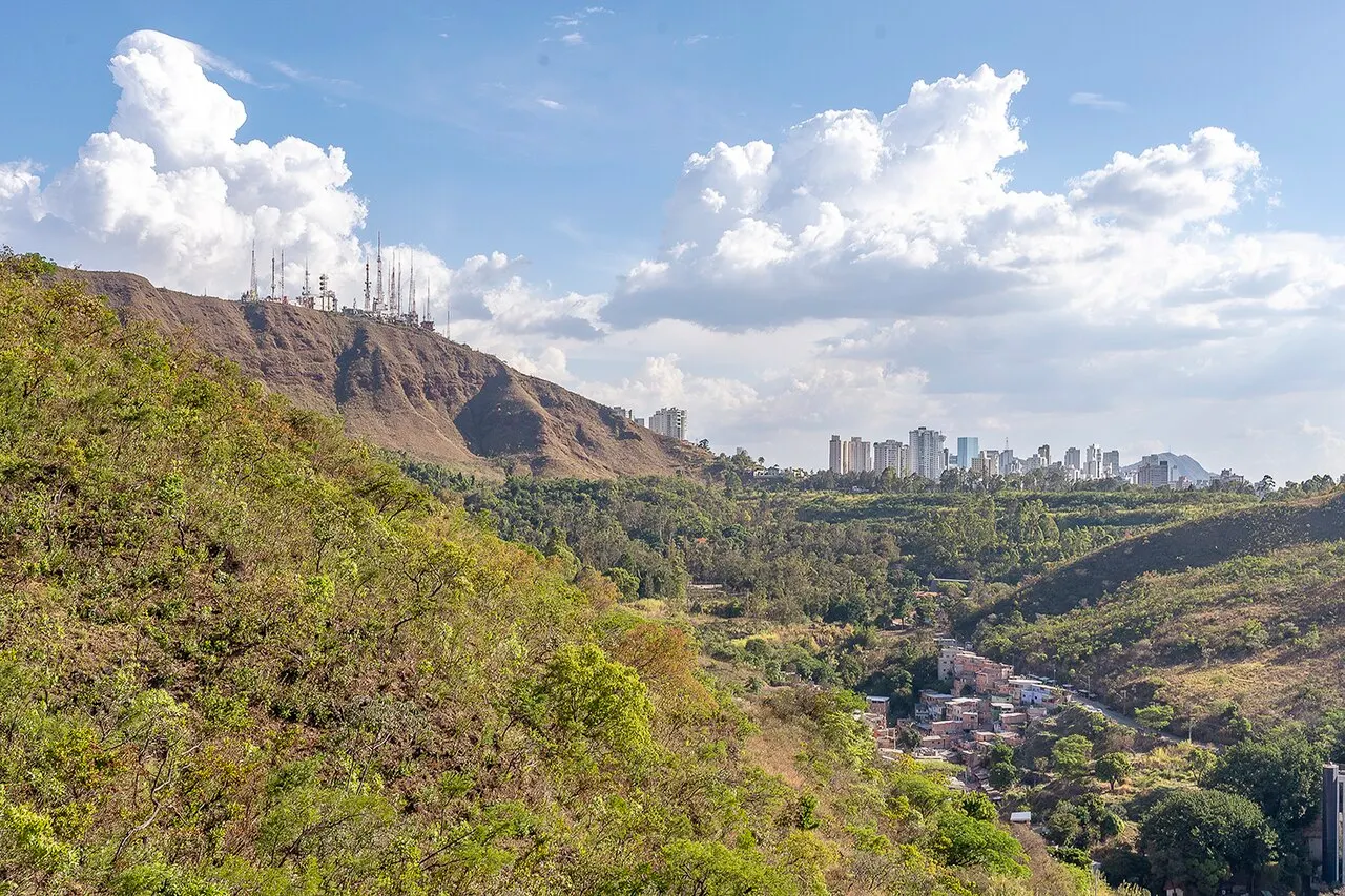 A encosta verde da Serra do Curral com as antenas no alto e os prédios da cidade ao fundo, sob nuvens brancas.
