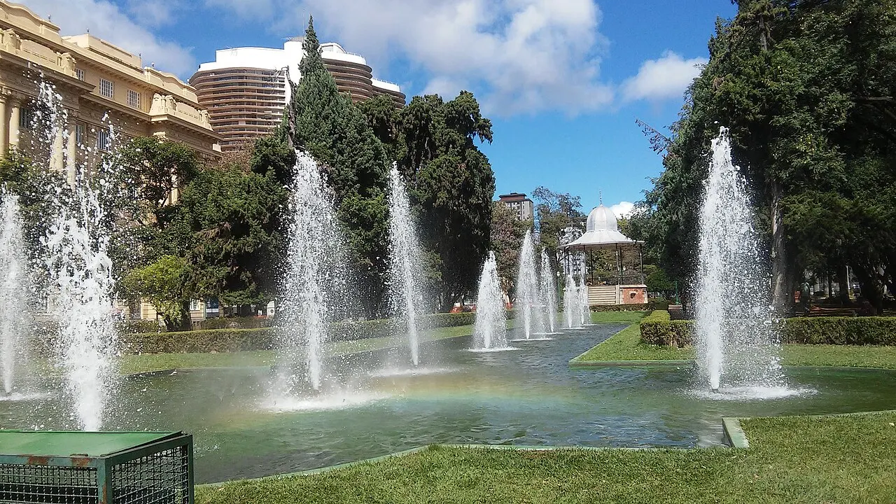 Os jatos da fonte da Praça da Liberdade sobre o espelho d'água, com o coreto e os prédios em volta.