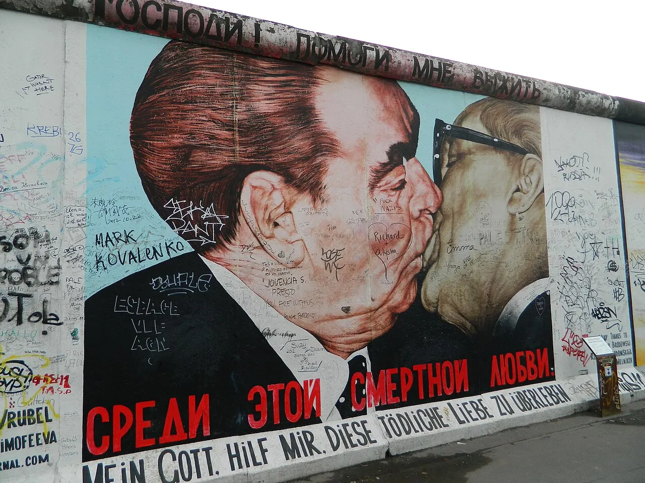 Trecho do muro da East Side Gallery com a pintura de dois homens se beijando e assinaturas por cima.