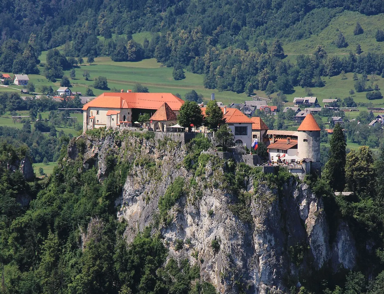 O Castelo de Bled, de telhados laranja, no alto de um penhasco de rocha clara sobre a floresta.