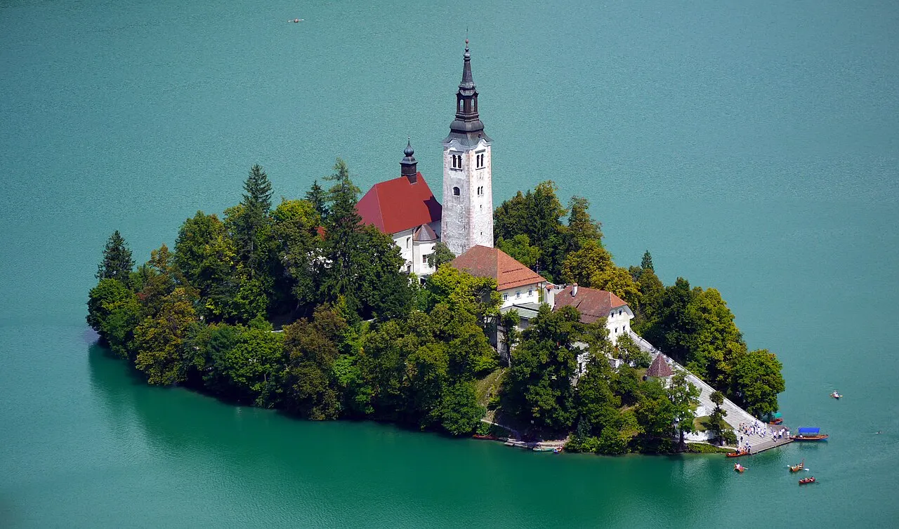 A ilha de Bled vista do alto, coberta de árvores, com a igreja de telhado vermelho, o campanário branco e a escadaria até a água.
