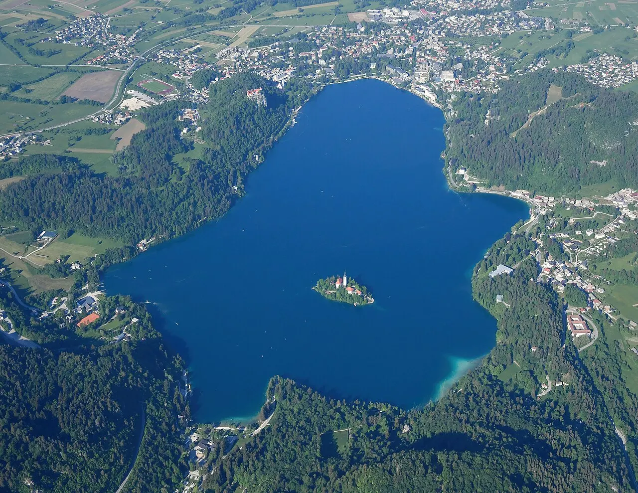 O Lago de Bled visto de avião, azul-escuro entre florestas, com a ilha pequena no meio e a cidade de Bled na margem.
