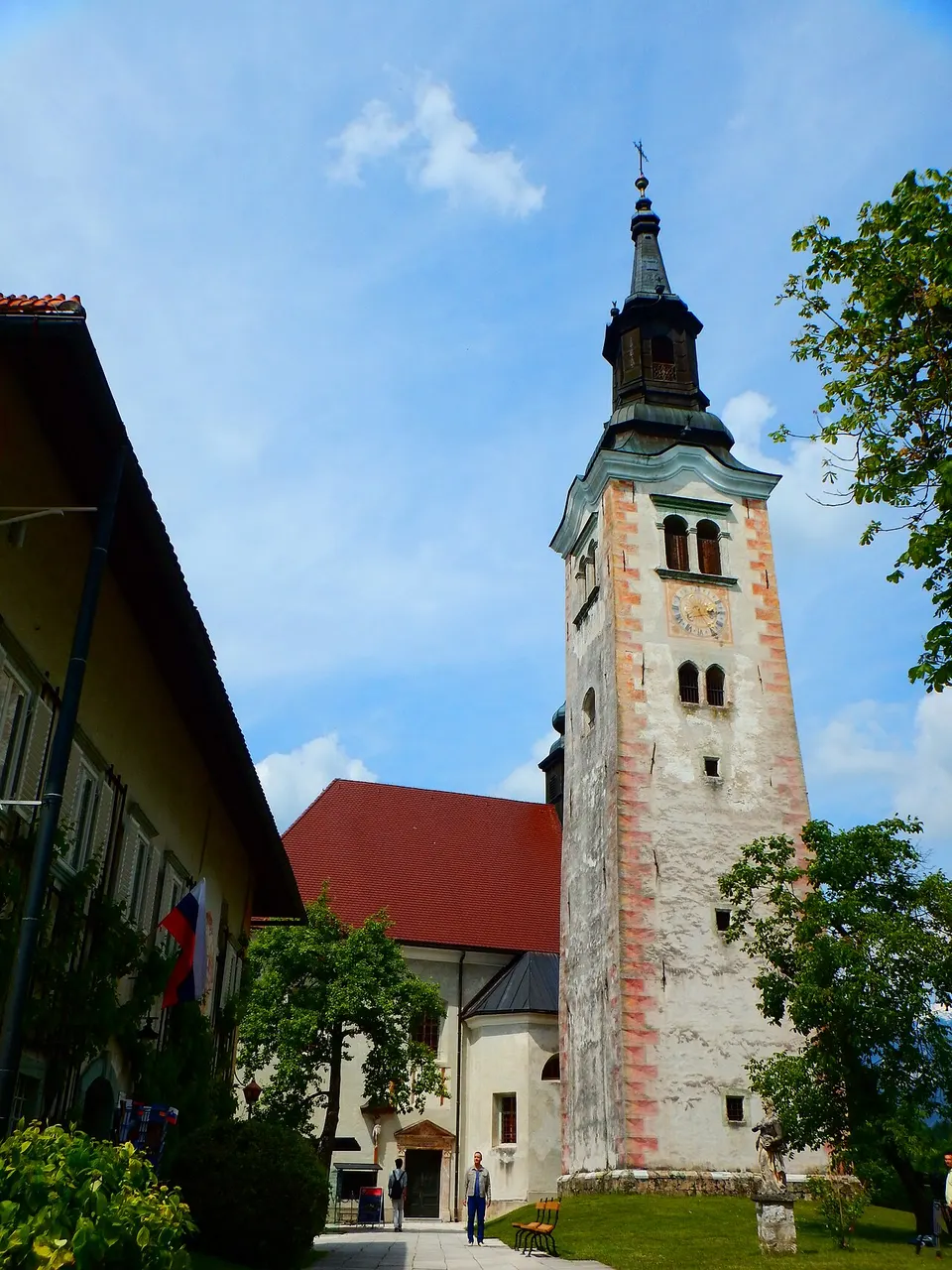 O campanário alto da Igreja da Assunção, na ilha de Bled, com a igreja de telhado vermelho e árvores em volta.