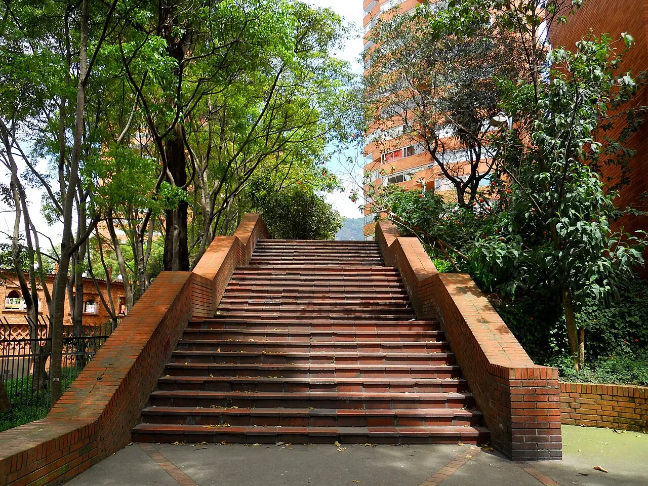 Ampla escadaria de tijolo com mureta curva sobe entre árvores até prédios residenciais de tijolo aparente, em dia ensolarado com sombras marcadas.