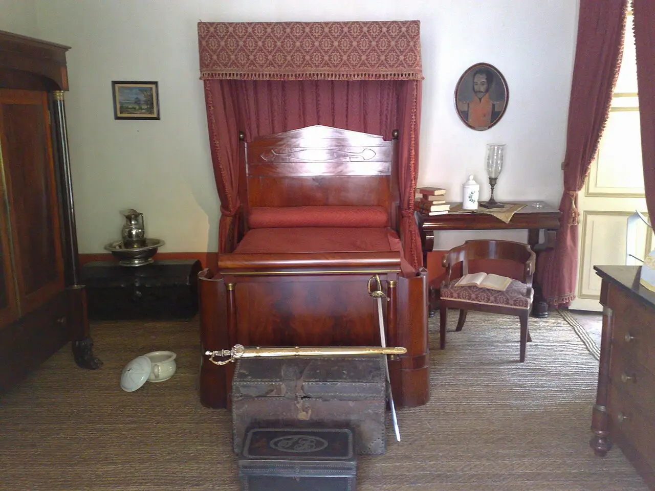 Quarto da Quinta de Bolívar, com cama de dossel vermelho, uma espada sobre um baú e um retrato na parede.