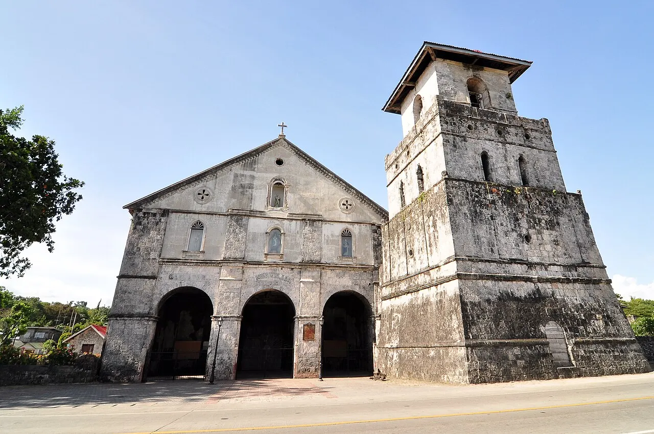 La iglesia de Baclayon, de bloques de piedra ennegrecidos, con tres arcos en la fachada y un campanario ancho y cuadrado al lado, junto a la calle.