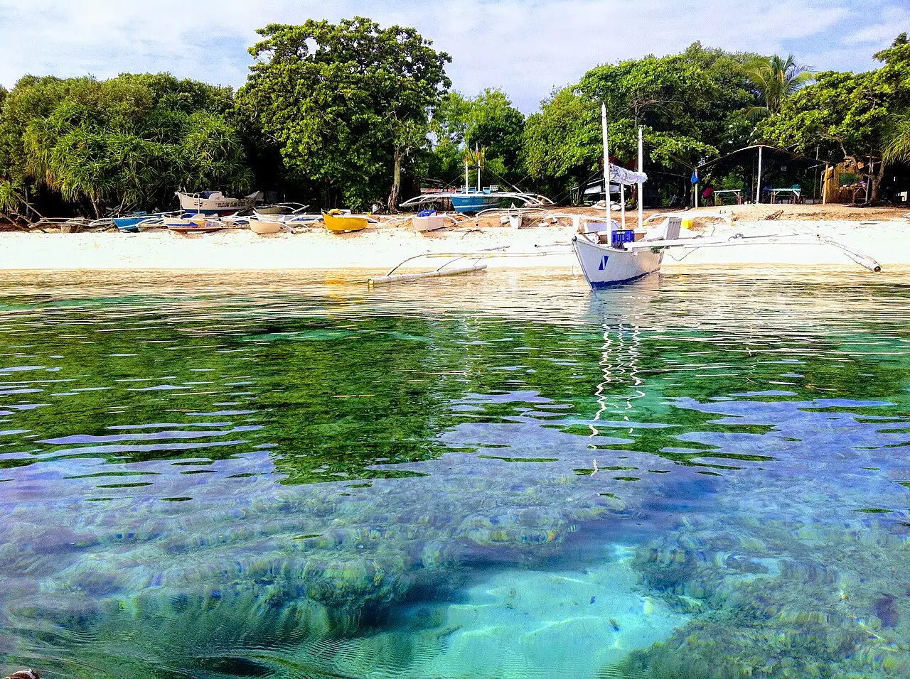 Agua poco profunda y transparente sobre el arrecife en primer plano; al fondo, una franja de arena blanca con barcas de balancines y árboles frondosos.