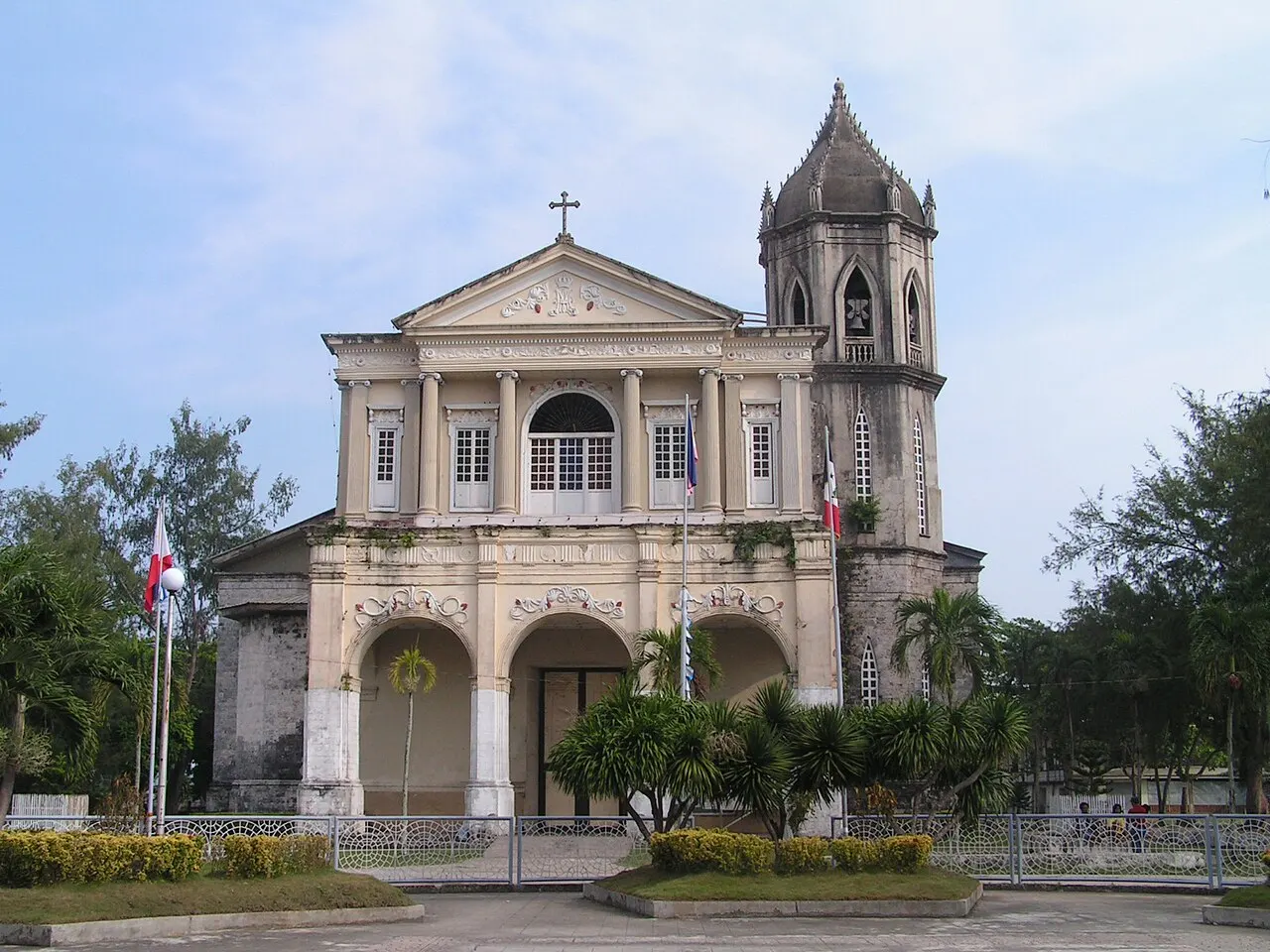 La iglesia de Dauis, de fachada clara con columnas, frontón triangular y arcos en la base, con un campanario puntiagudo al lado, tras un jardín vallado.