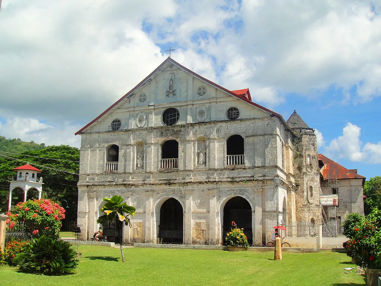 La fachada de piedra clara y manchada de la iglesia de Loboc, con tres arcos en la base y hornacinas con estatuas, ante un césped con arbustos en flor.