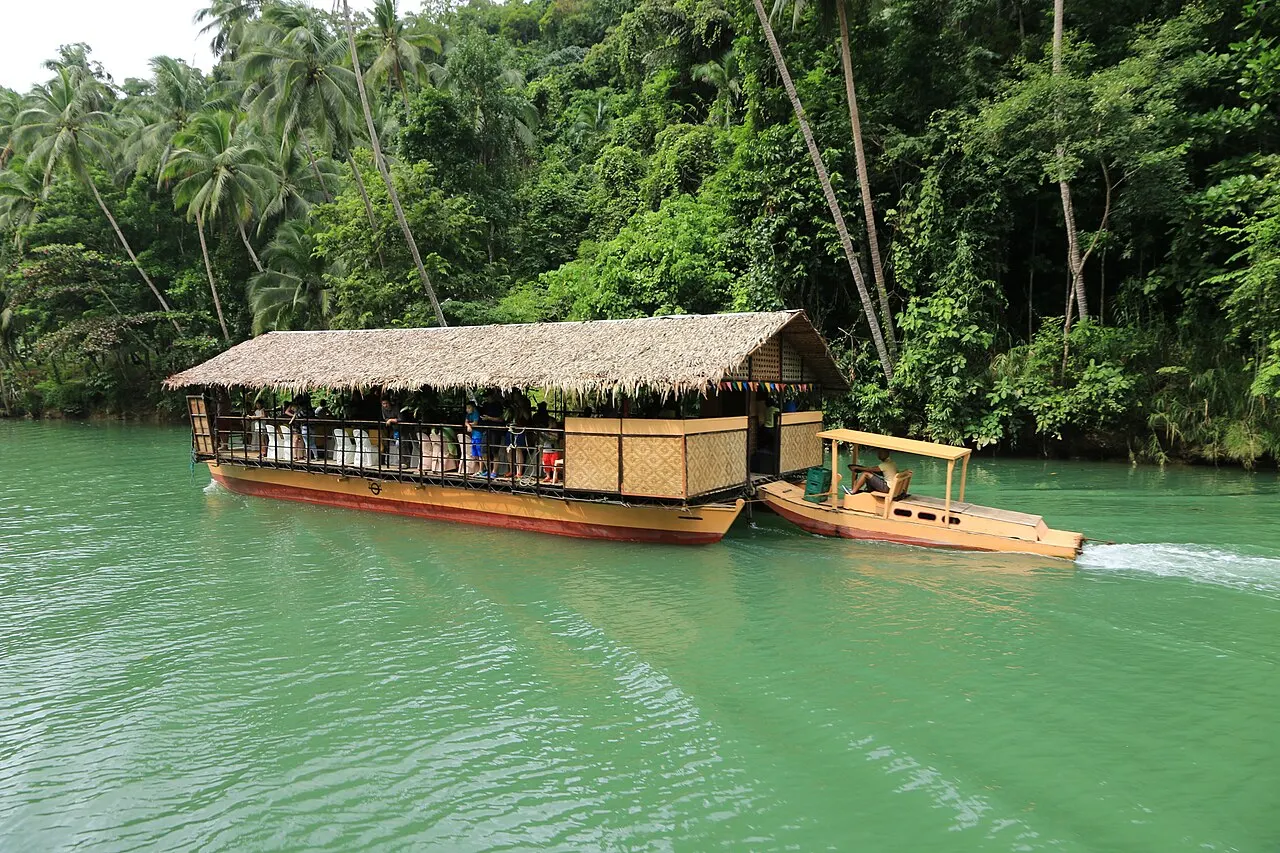 Una embarcación ancha con techo de paja, remolcada por otra más pequeña, baja por el río de agua verde entre orillas de selva densa y cocoteros.