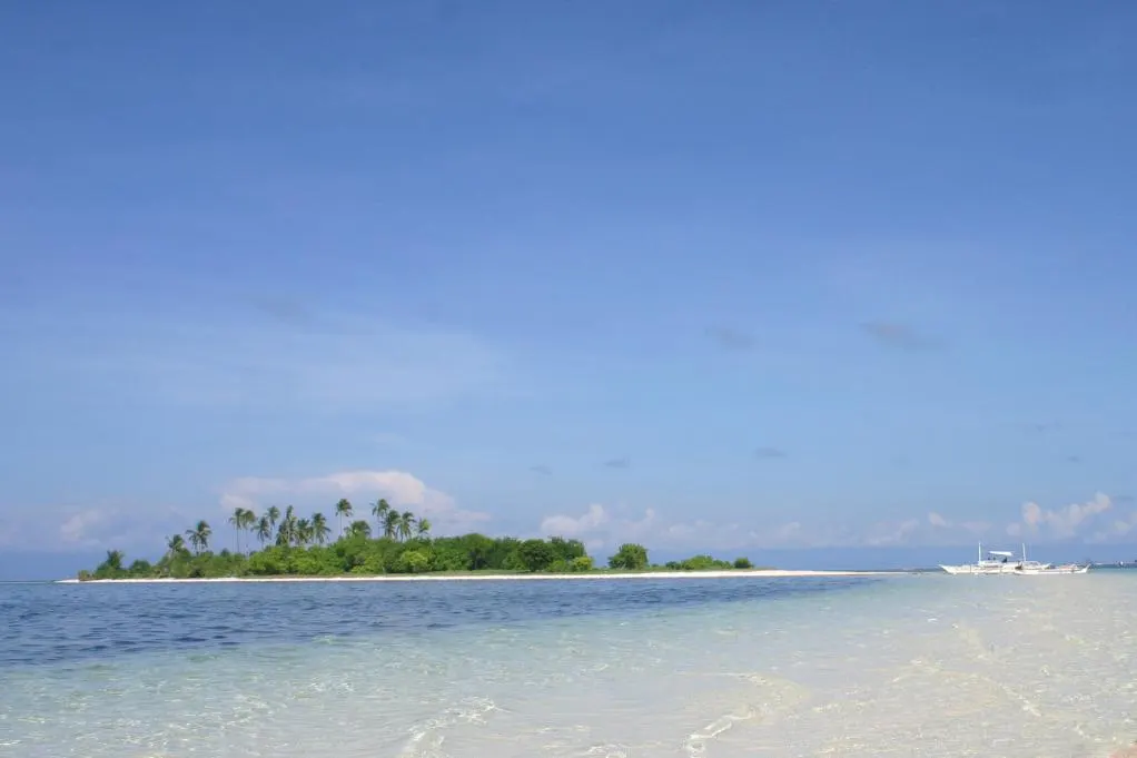 Agua clara y poco profunda sobre un banco de arena en primer plano; al fondo, un islote bajo con cocoteros y una barca de balancines bajo el cielo azul.