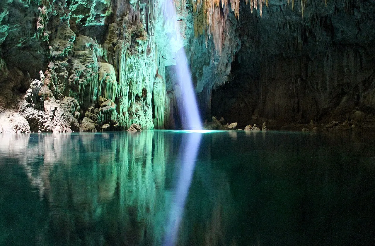 Um raio de luz descendo da abertura do Abismo Anhumas até o lago escuro e verde, com formações de calcário refletidas na água.