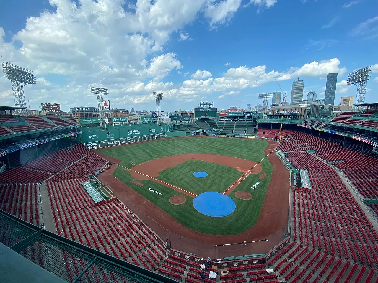 O campo de beisebol do Fenway Park está vazio, com as arquibancadas vermelhas, o muro verde e o centro de Boston ao fundo.